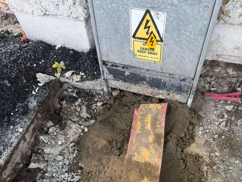 An excavation beside a feeder pillar with a "Caution electric" cable cover board exposed in the ground.