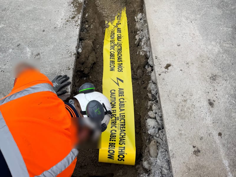 An operative in a hard hat, ear defenders, gloves and hi-vis laying yellow "Caution electric cable below" warning tape in a trench.