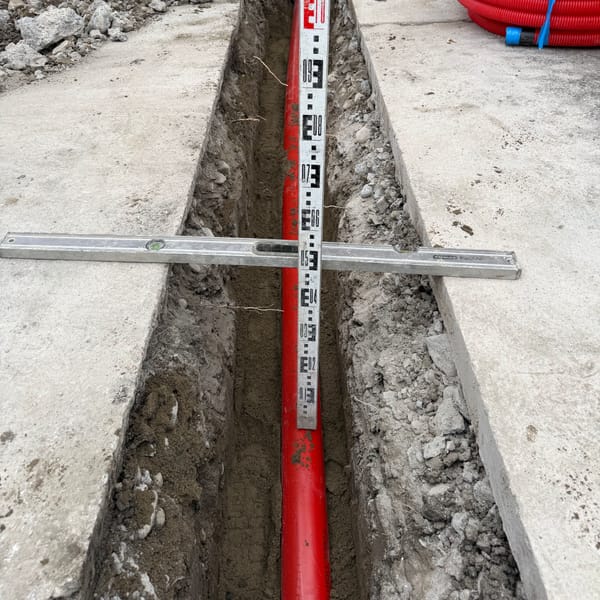 A level staff and straightedge across a narrow trench, recording the depth of the red duct below.