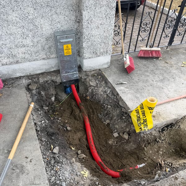 A red duct running from a mini-pillar through an open excavation, with cable warning tape at the edge.