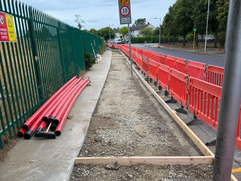 Red duct lengths stacked beside a footway prepared with formwork, behind barriers.
