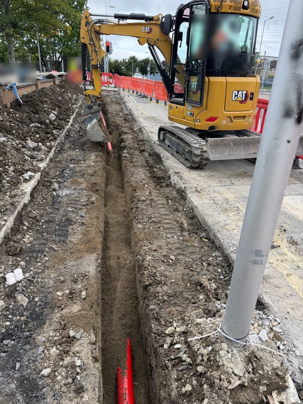 A CAT mini excavator digging a duct trench along a footway, with pedestrian barriers along the road edge.