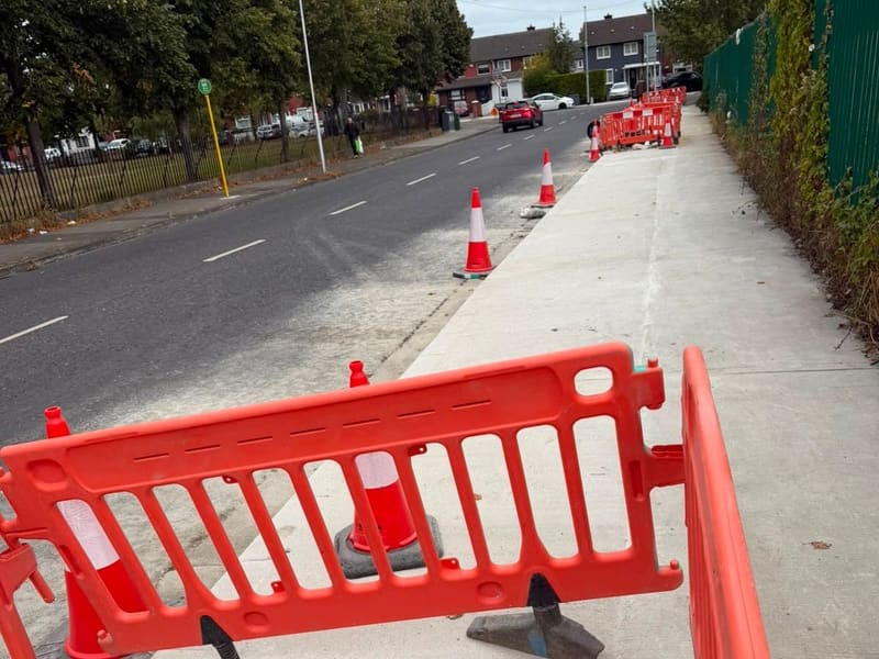 Finished concrete footway beside a main road, with barriers and cones still in place.