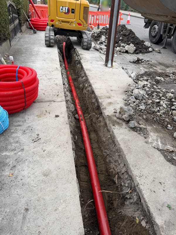 A long open trench in a concrete footway with red duct laid along the bottom, a coil of duct beside it and a mini excavator behind.
